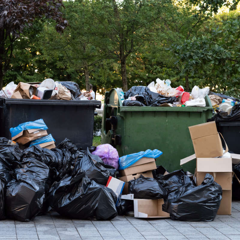 Das Bild zeigt mehrere Müllcontainer mit je 1.100 Liter Fassungsvermögen welche stark überfüllt zur Abholung bereitstehen. Davor stehen zusätzlich zahlreiche schwarze Müllsäcke.